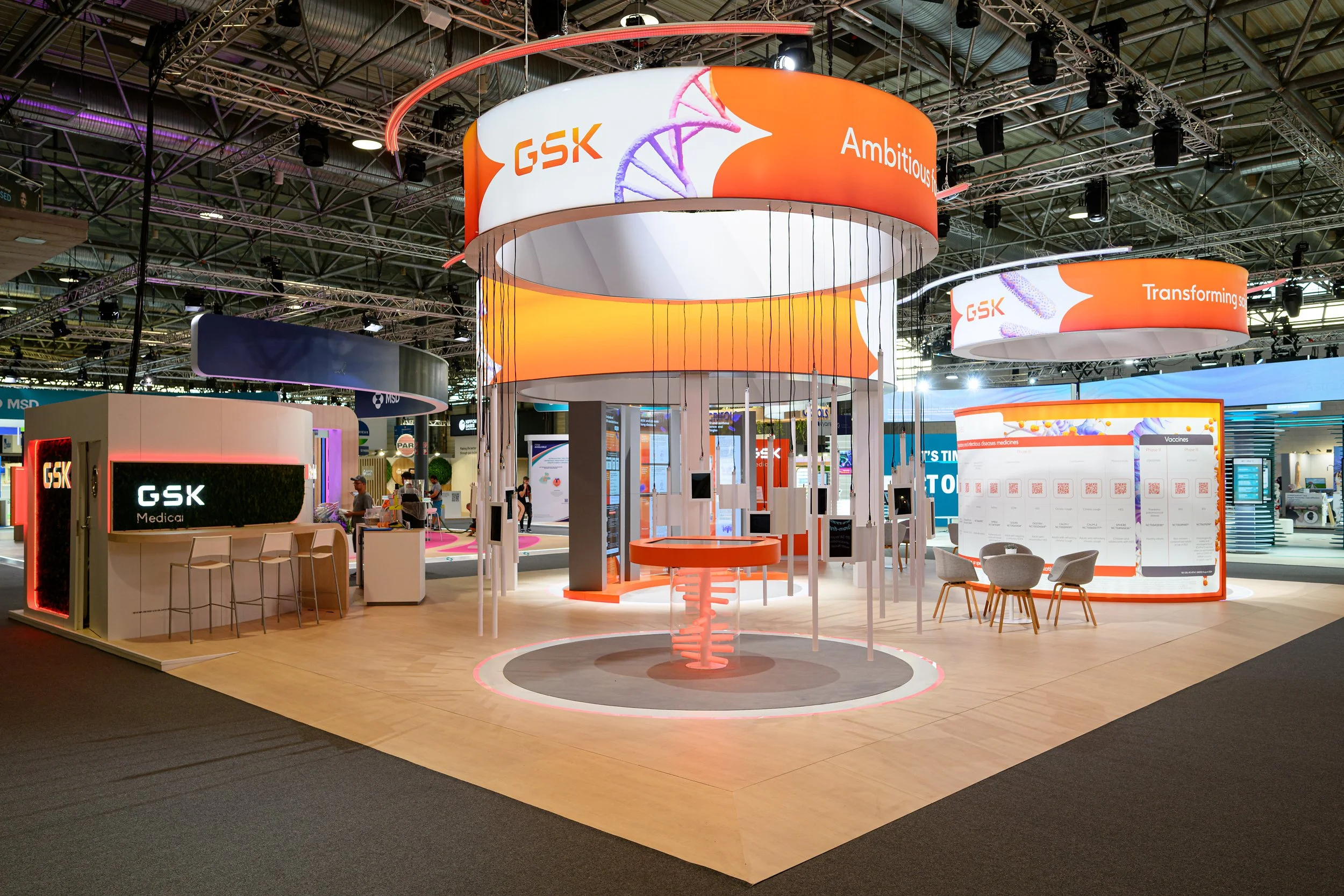 Exhibition booth with GSK branding, including circular hanging banners with orange and white colors, display screens, and seating areas at a trade show or conference.