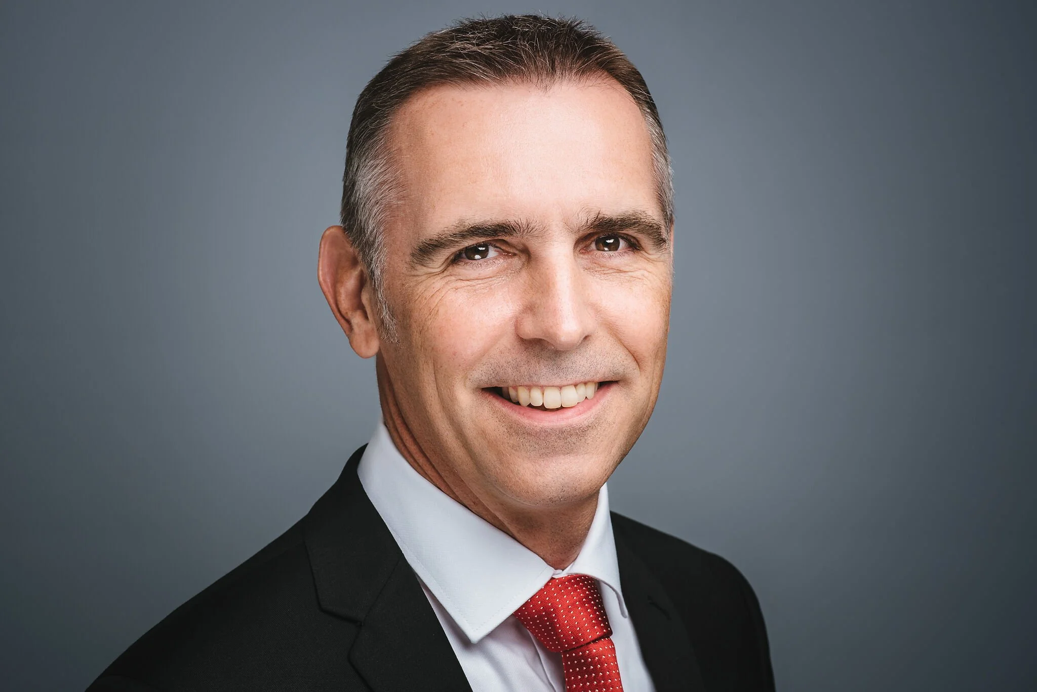 A professional headshot of a smiling man in a suit and red tie against a gray background.