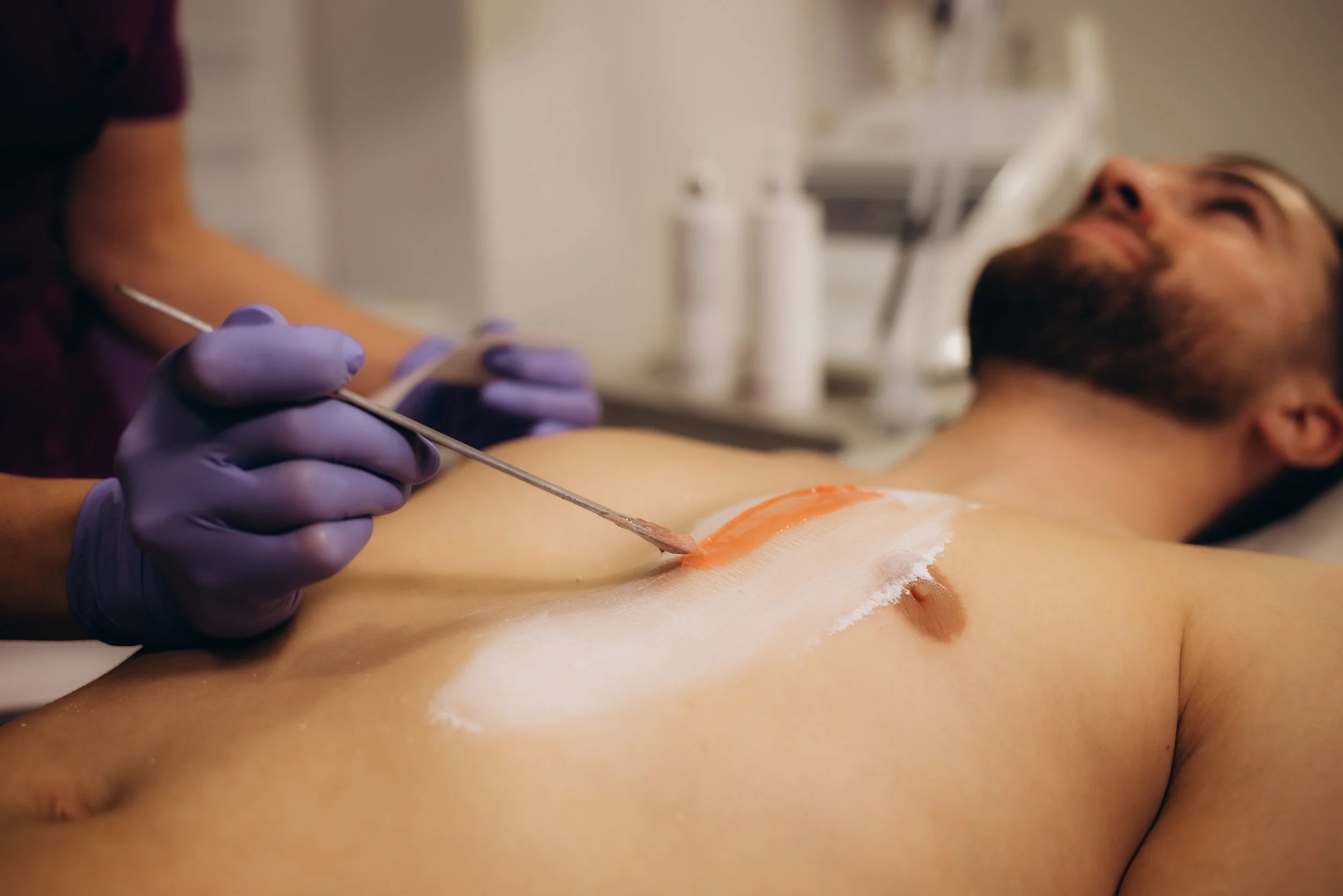 An expert in purple gloves applies wax to a man's stomach with a spatula during a body waxing treatment.