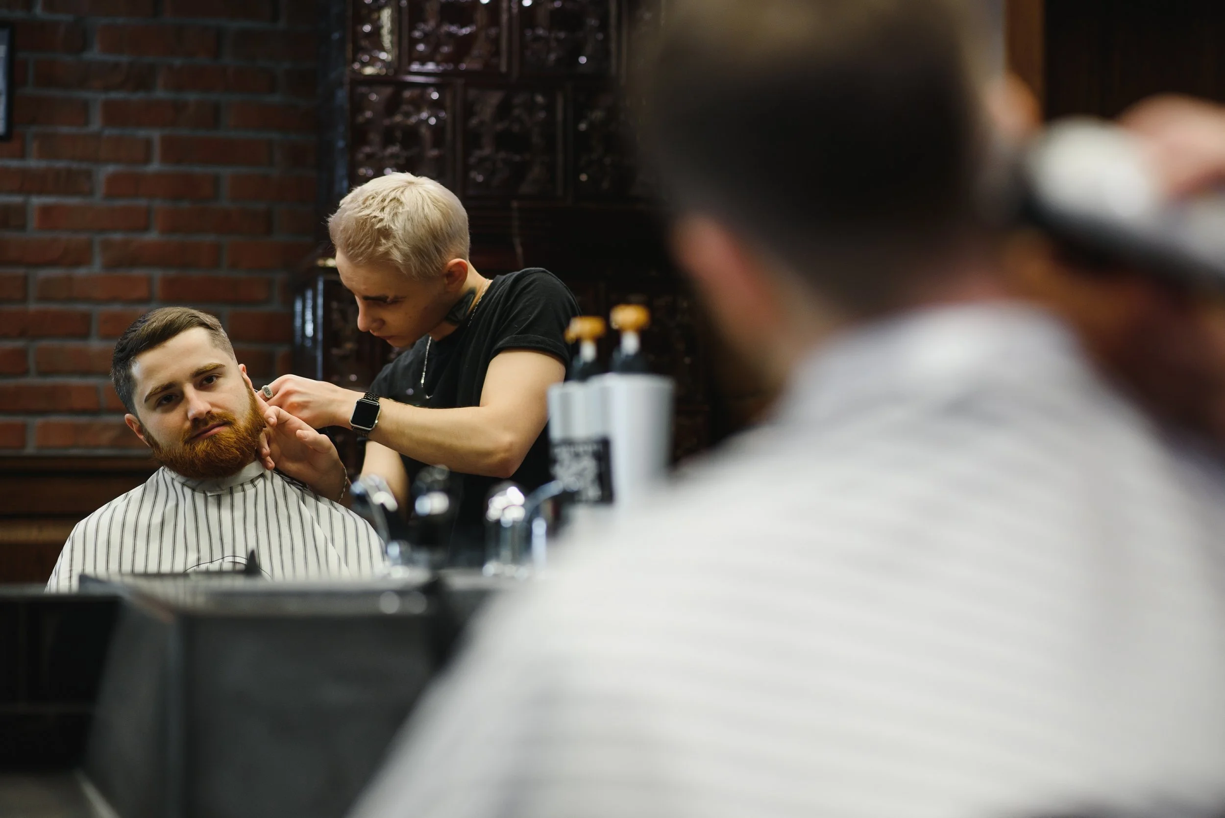 A barber trims a seated client's beard by an exposed brick wall during a men's grooming visit in Chicago.
