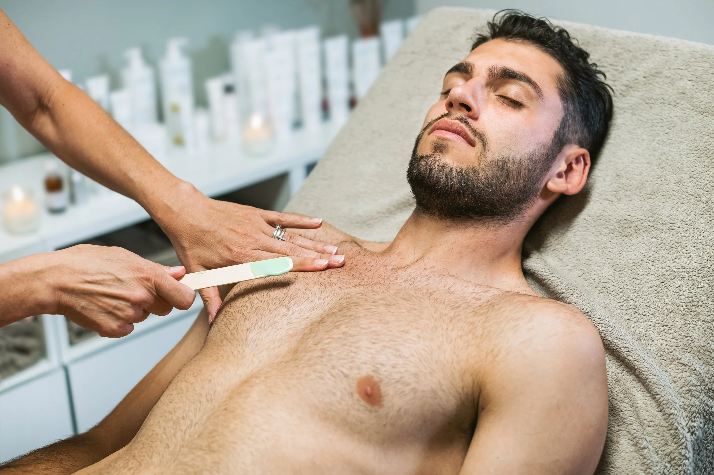 An esthetician in white gloves presses a wax strip onto a man's lower abdomen during a body waxing session.