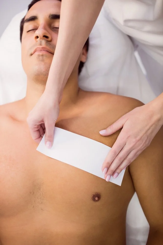 Technician removing a wax strip from a male client's chest during a professional mens waxing session in a salon treatment room.