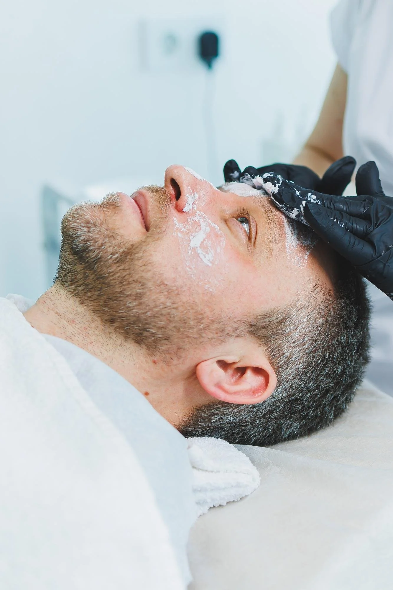 A man with a short beard is lying on a treatment bed while a practitioner applies a skincare product to his forehead during a clinic procedure, discussing sun exposure after a chemical peel.