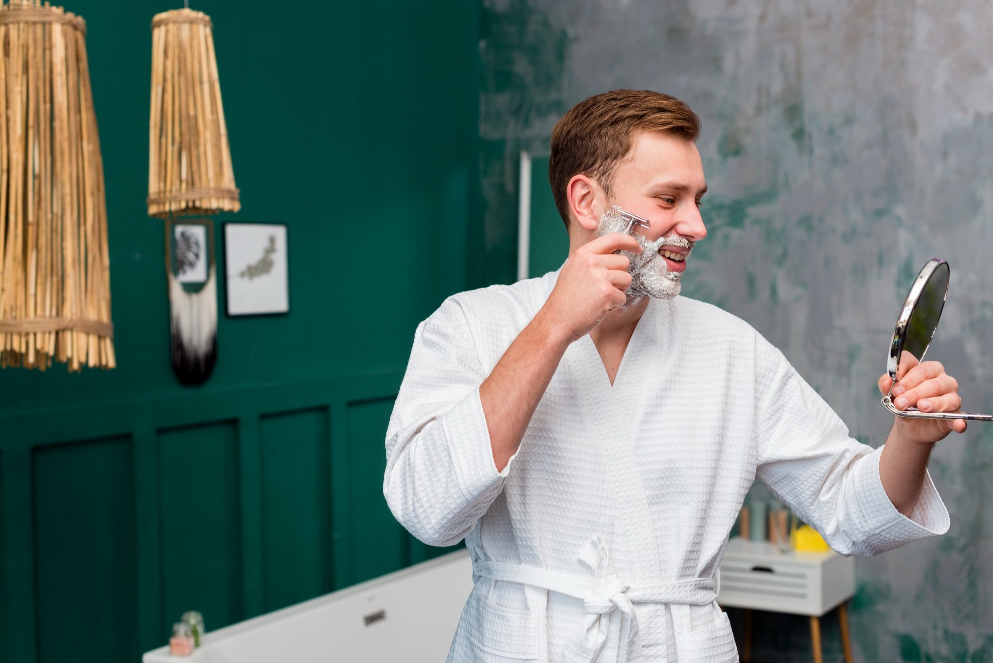 Man in a white robe shaving his face with a razor while holding a mirror in a modern bathroom during his at-home grooming routine.