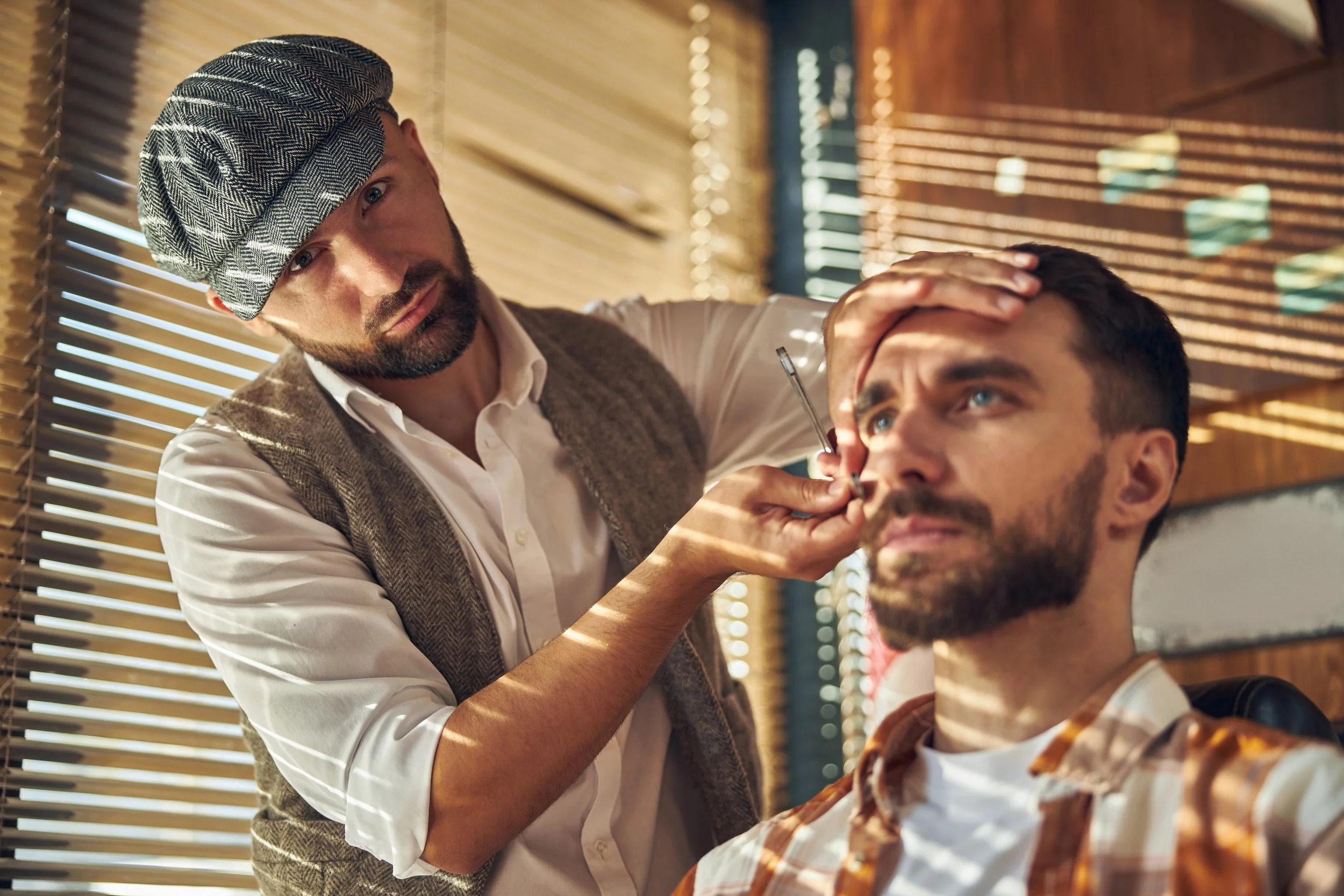 A barber in a flat cap grooms a client's beard by a sunlit window at a men's salon in Chicago.