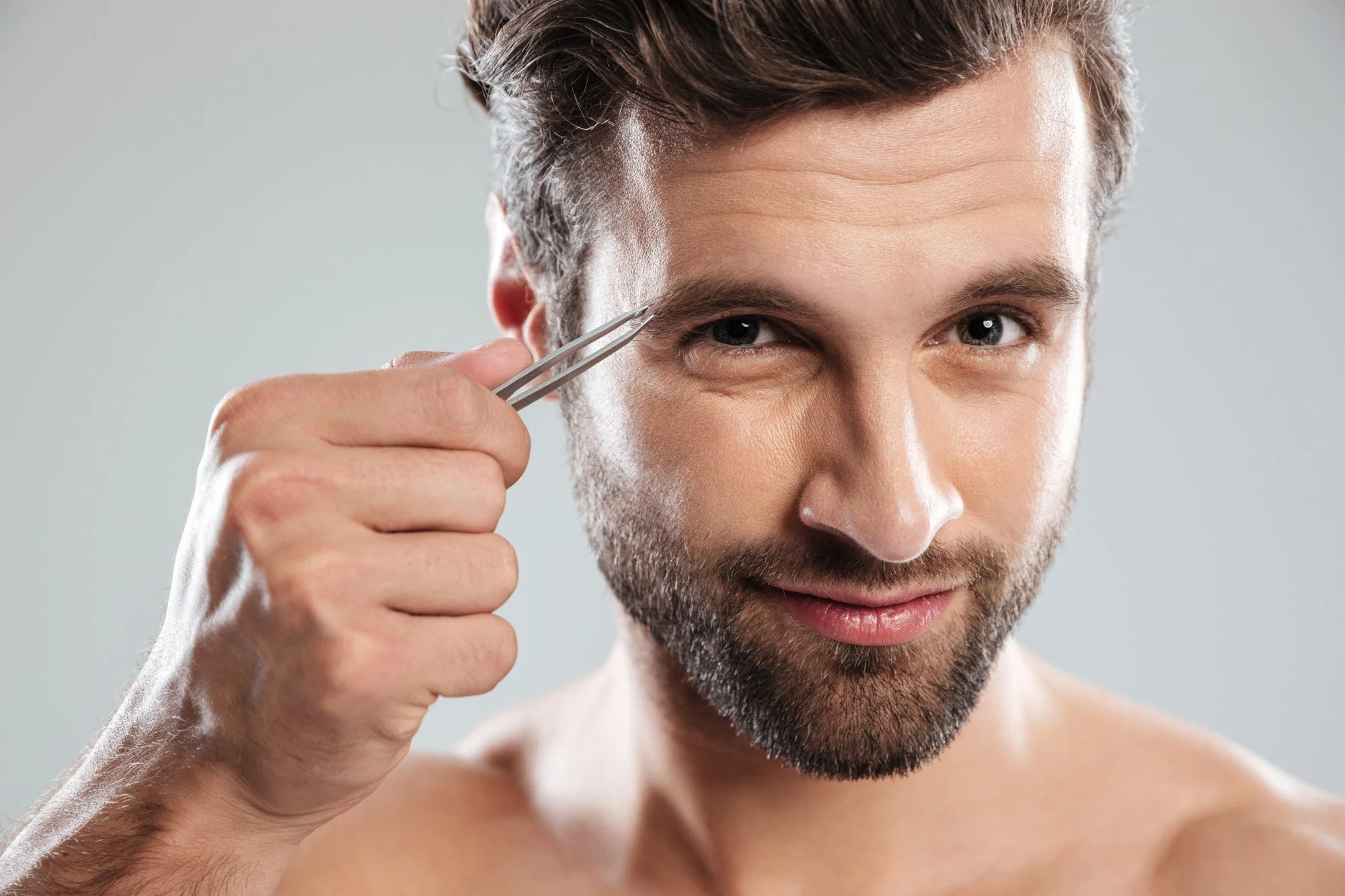 Man using tweezers to spot-pluck stray hairs as part of at-home men's eyebrow grooming.