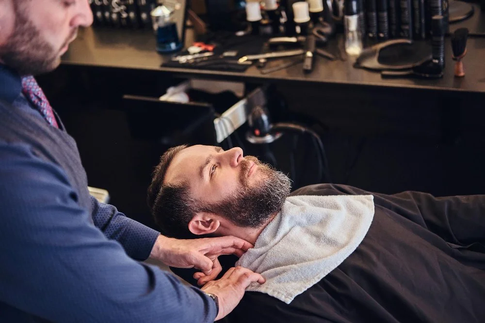 A master barber carefully prepares a male client's face with shaving lather before a professional straight razor shave in a barbershop.