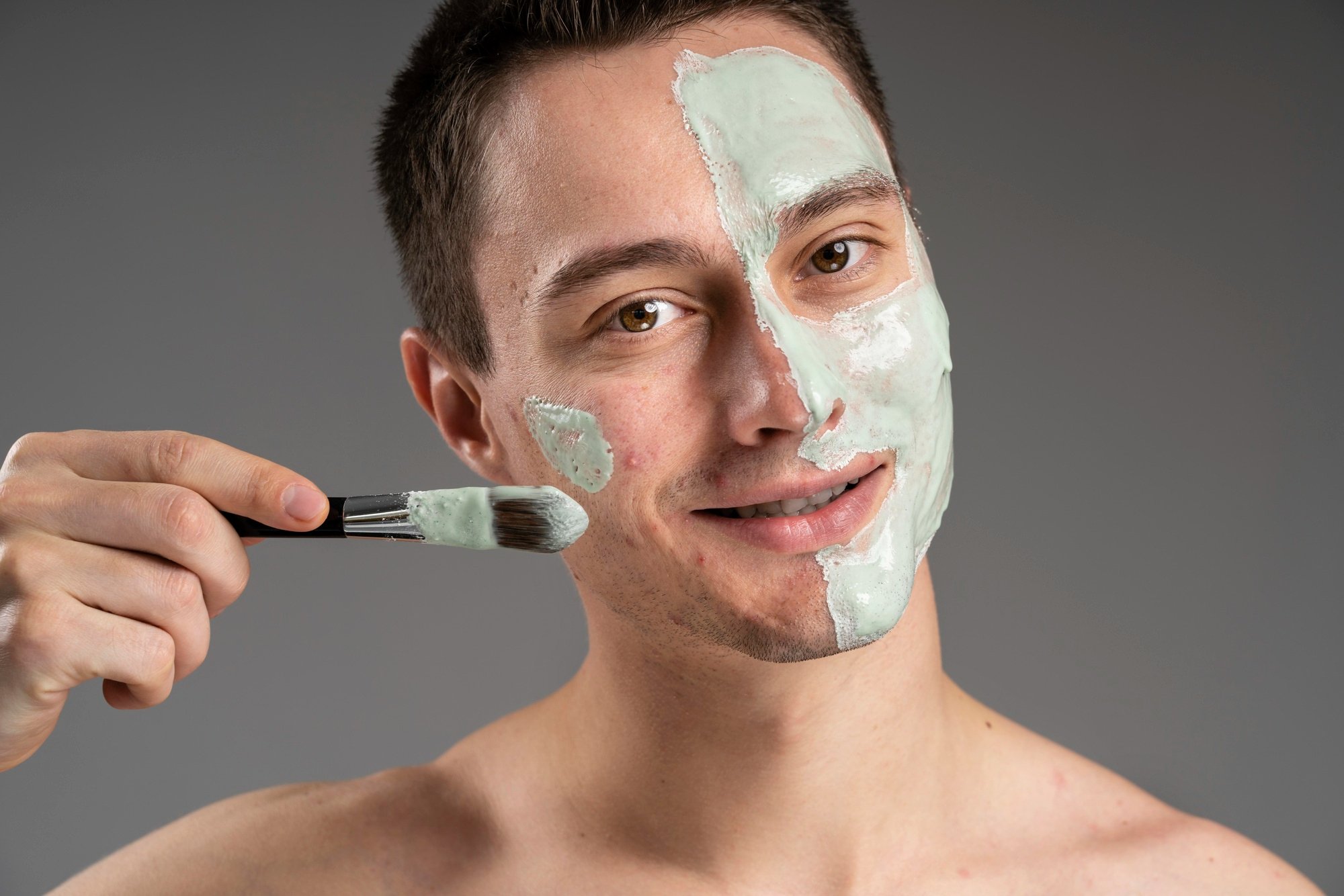 Man smiling as a clay facial mask is brushed onto half of his face, showing the treatment in progress against a grey backdrop.