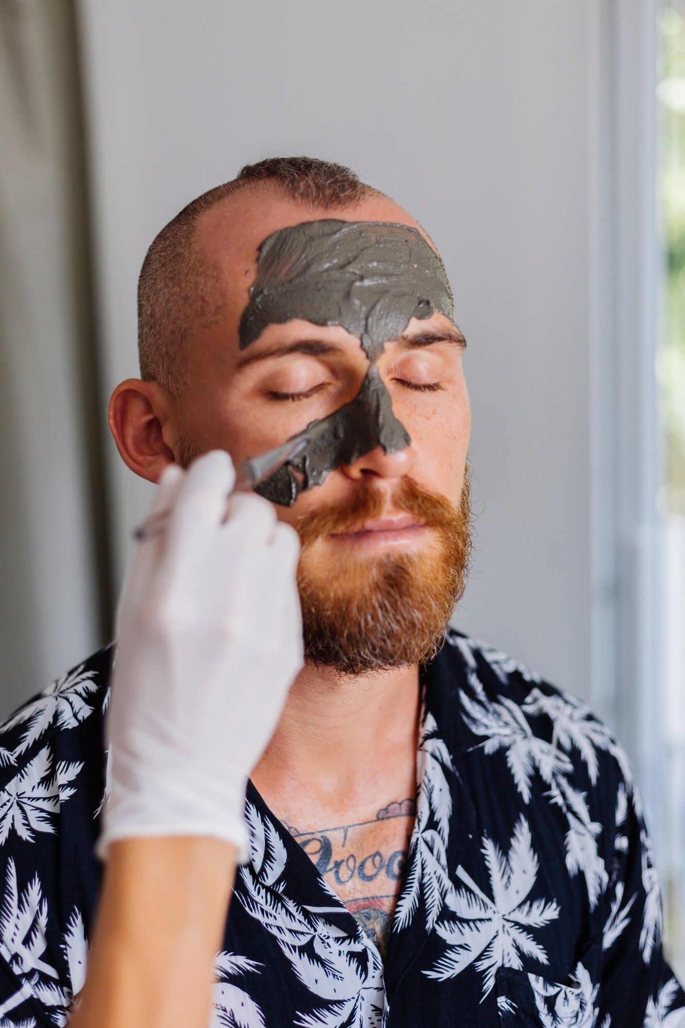 Skincare professional applying a dark facial mask to a bearded man’s face while he sits with eyes closed, illustrating options for facial treatments and the best time for a chemical peel.