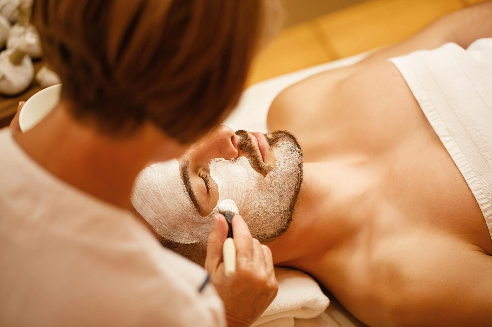 Esthetician applying a white facial mask to a man's face with a brush while he lies down during a professional facial treatment.