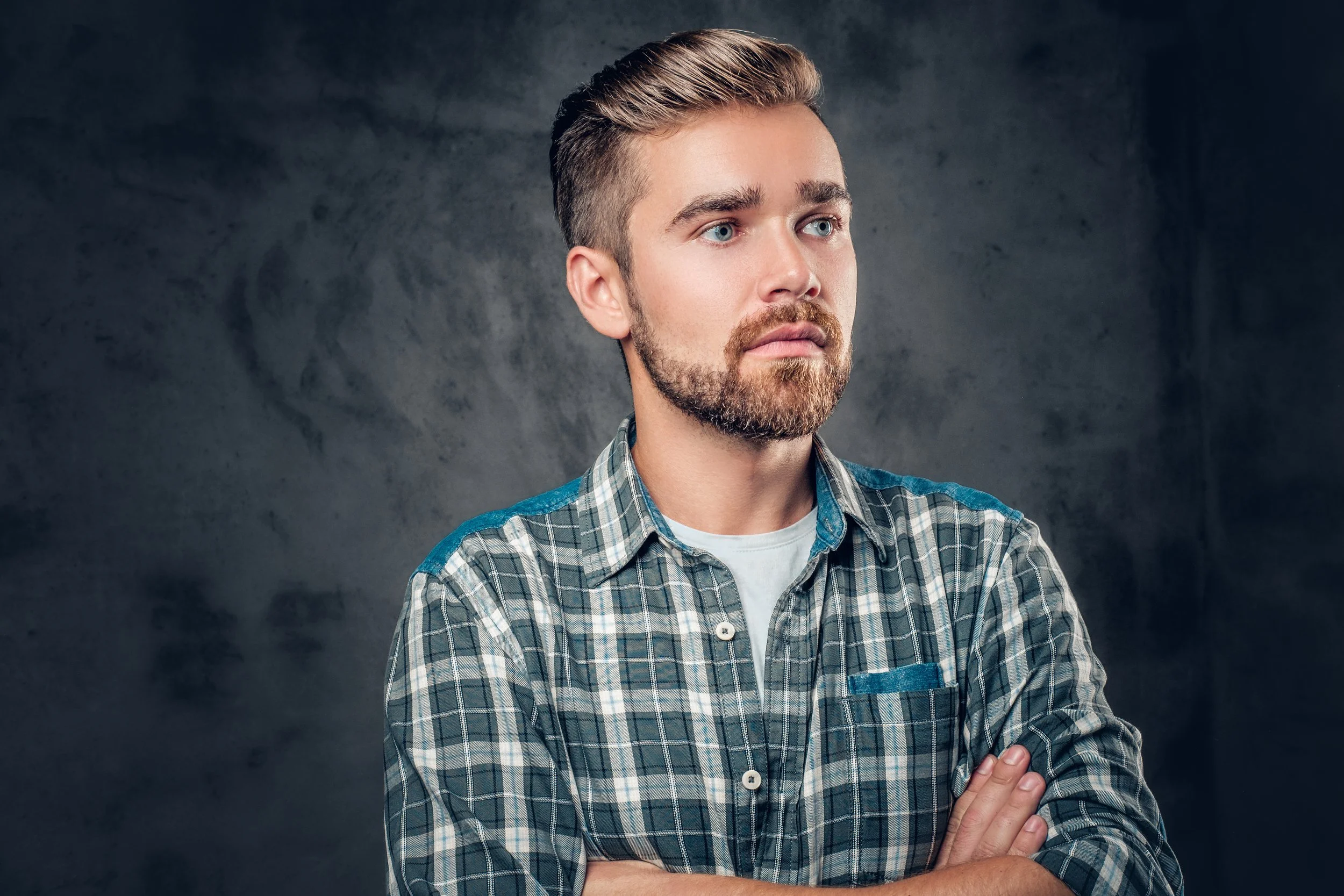 Man with a straight, textured crop haircut and full beard, wearing a plaid shirt with arms crossed against a grey studio backdrop.