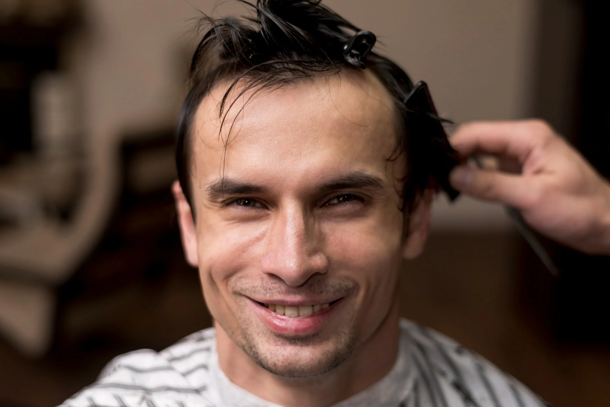 Smiling man getting his hair sectioned and clipped by a barber during a haircut appointment, with damp hair styled at the front.