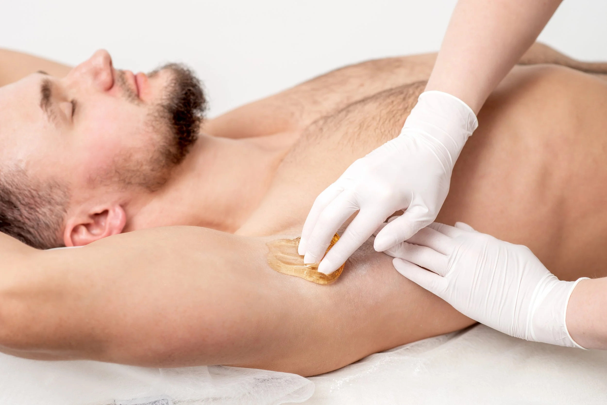 A gloved therapist applies wax to a man's underarm with a spatula during an underarm waxing treatment.