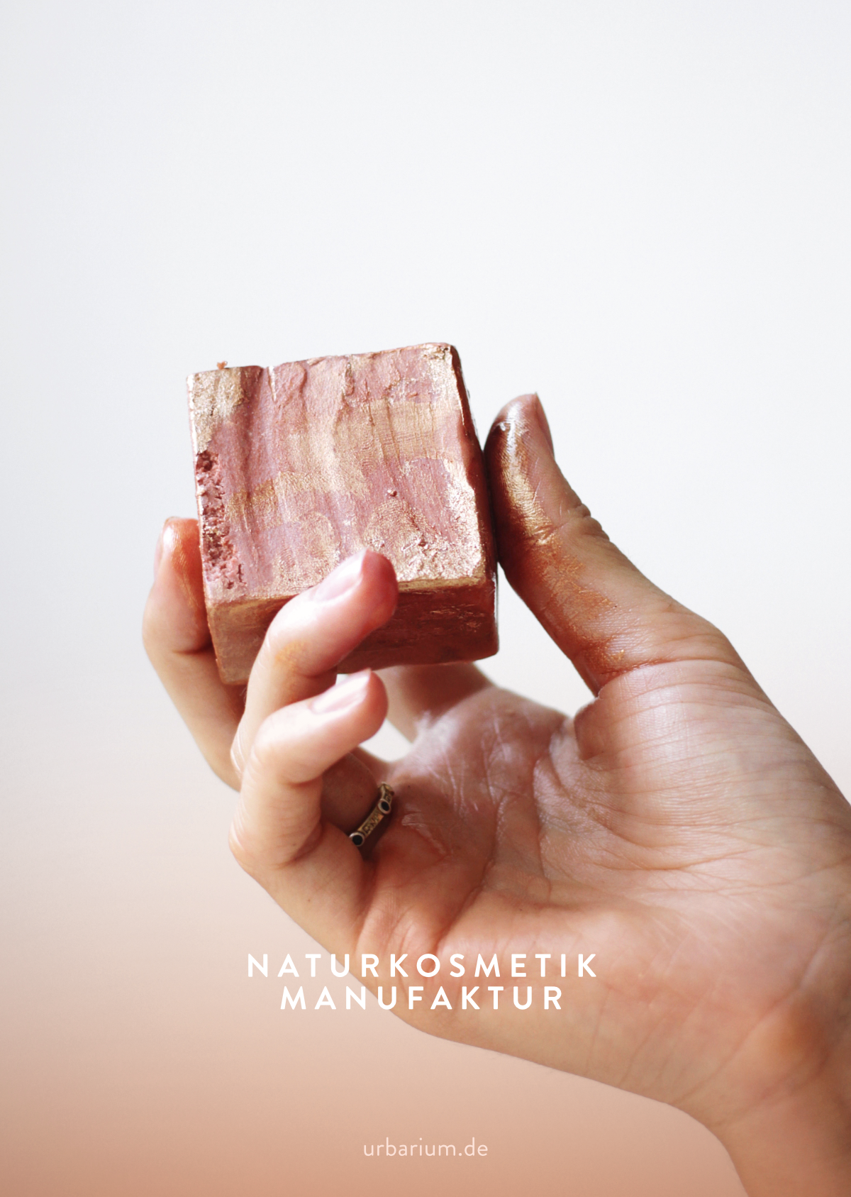 Eine Hand hält eine natürliche Kosmetikseife mit goldenen Akzenten, Text: 'Naturkosmetik Manufaktur, urbarium.de' im Hintergrund.