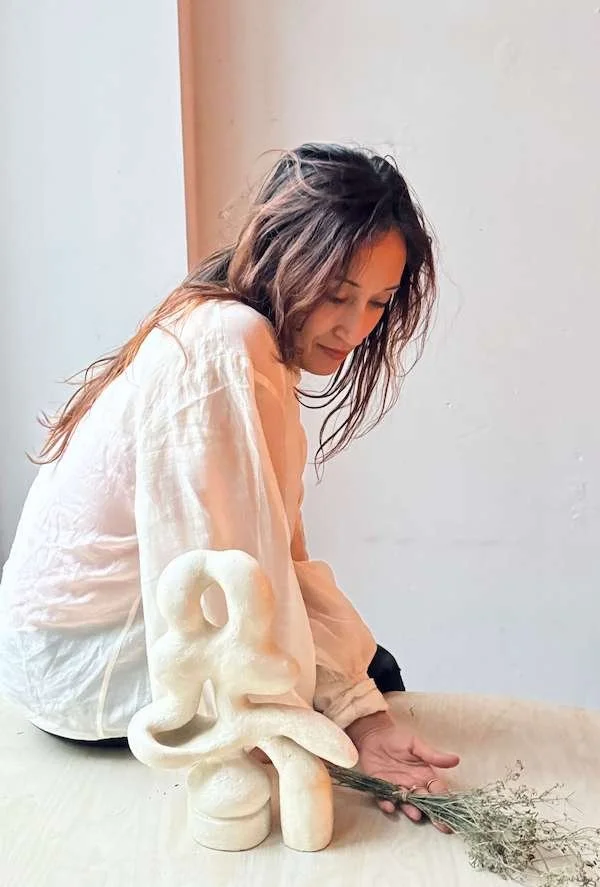 Eine Frau mit braunen, zerzausten Haaren in einem weißen, durchscheinenden Hemd sitzt an einem Tisch, in der Nähe befindet sich eine moderne Skulptur aus hellem Material und ein kleines Bündel getrockneter Kräuter oder Blumen.