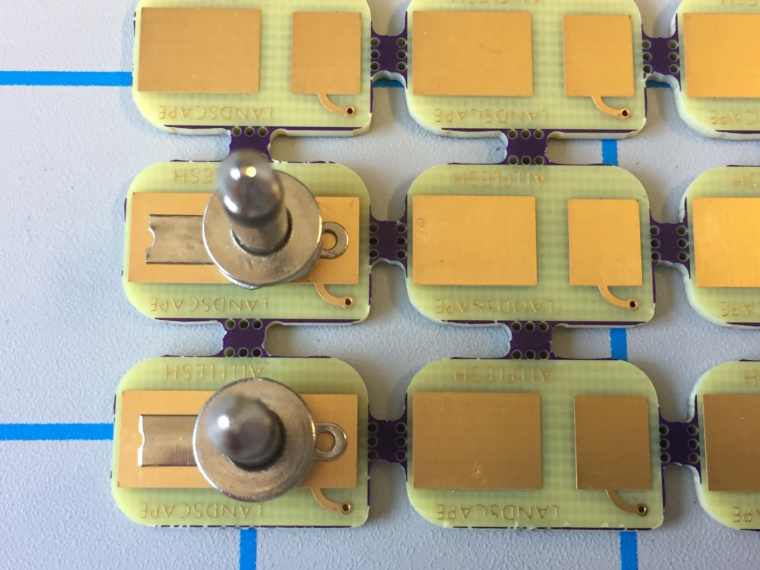 5.  Place your plugs atop the pcb surface as pictured so that the  center of the plug is in the space between the two large golden solder pads . Your plug should form a "T" shape standing up straight and perpendicular to the surface in both directions. The bent lugs of the plug should be flat against the gold solder pads with no airspace beneath them. Use your pliers to further flatten and adjust them at this step if needed.