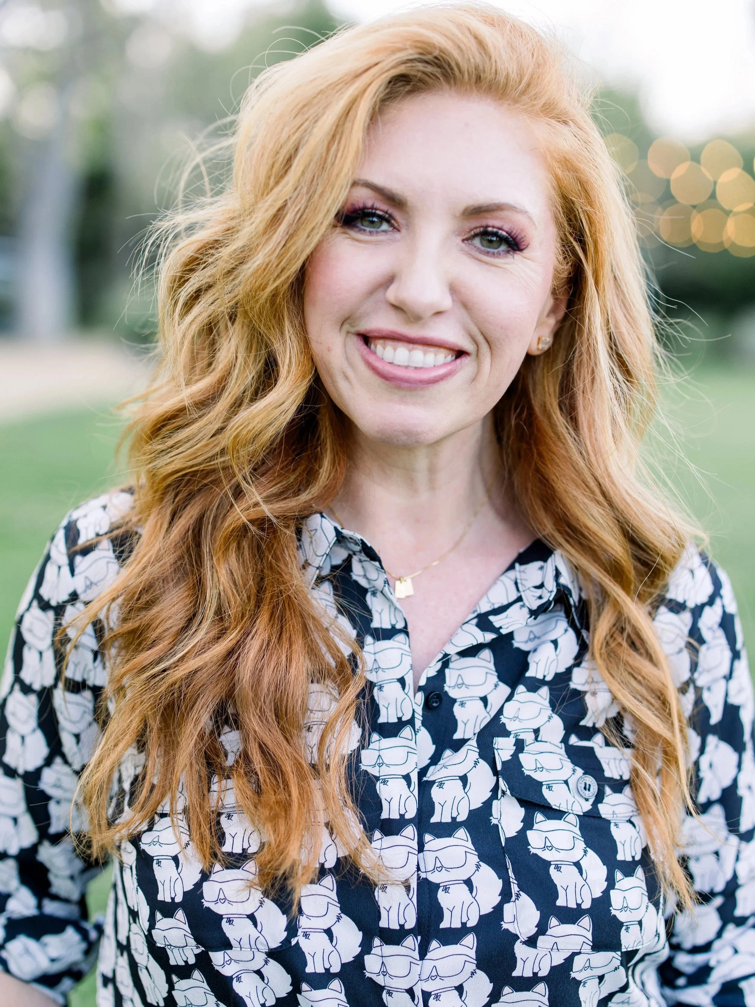 A woman with long red hair, blue eyes, and a bright smile, wearing a black and white patterned shirt with a dog motif, standing outdoors with blurred background of trees and Make-A-Wish lights.