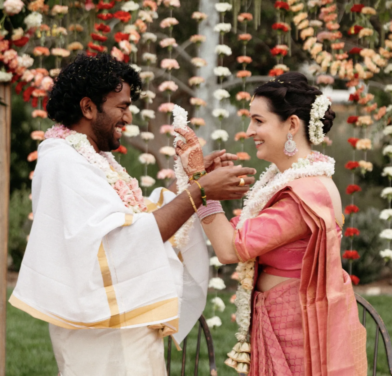 A couple celebrating a wedding outdoors, exchanging garlands and smiling, with floral decorations in the background.