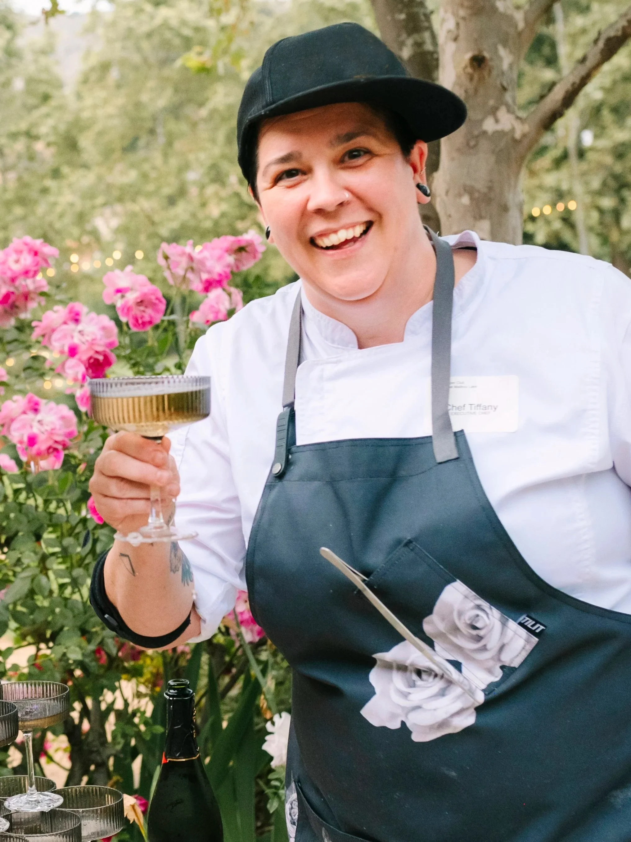 A smiling person with short dark hair, wearing a black cap, black earrings, a white shirt with a name tag, and a black apron, holding a wine glass outdoors near pink flowers and green trees.