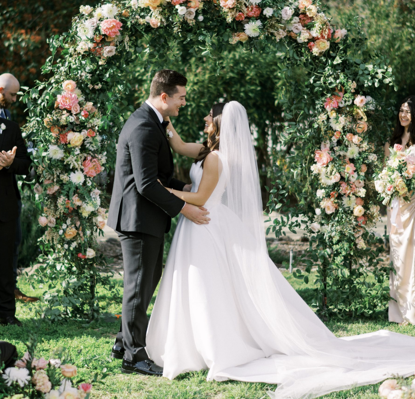 A newlywed couple stands together, smiling, during their outdoor wedding ceremony under a floral arch, with the bride in a white gown and veil and the groom in a black tuxedo, surrounded by friends and family.
