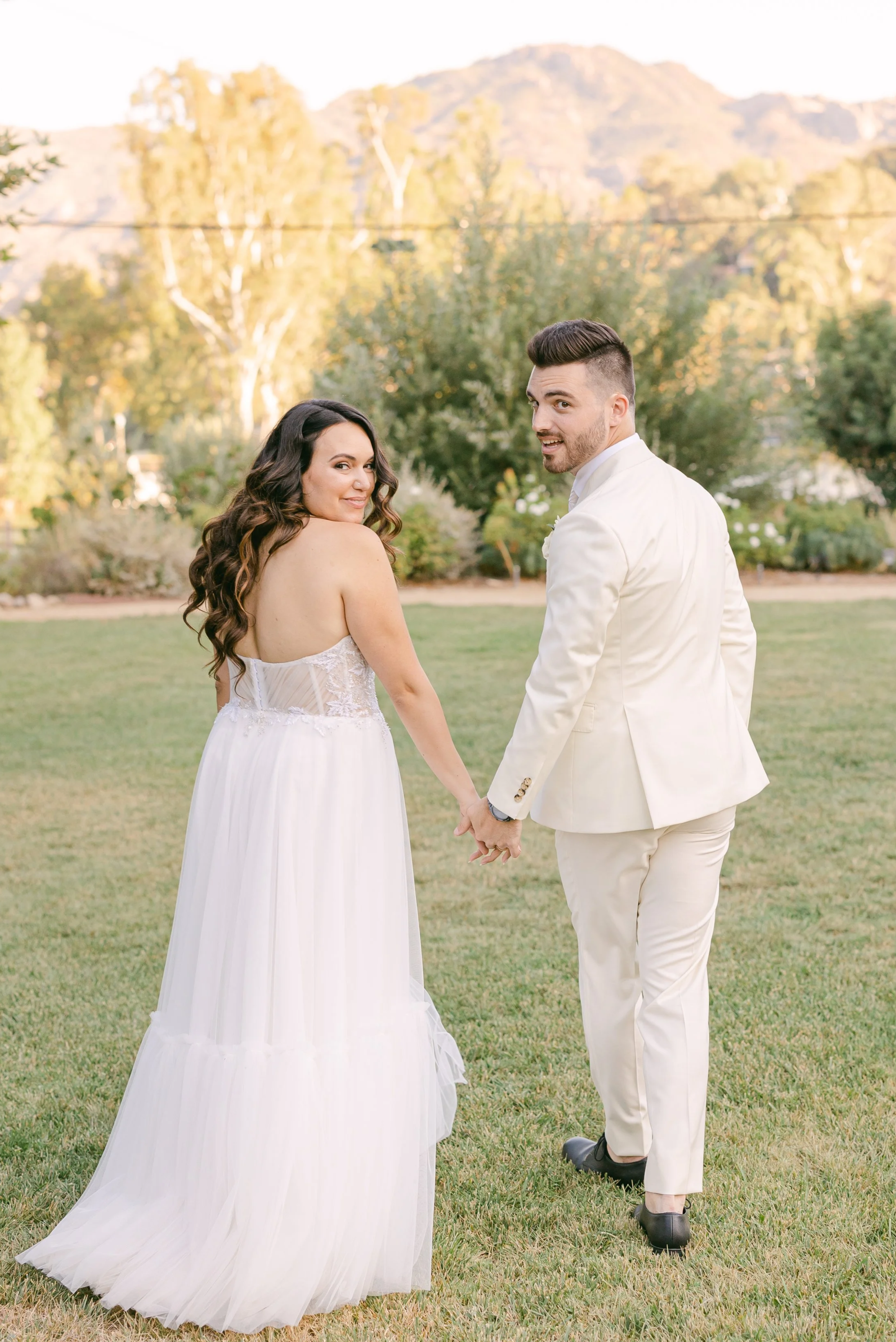 A bride in a white dress and a groom in a white suit hold hands looking over their shoulders to camera, at their summer wedding at The Lodge at Malibou Lake near Ventura County CA.