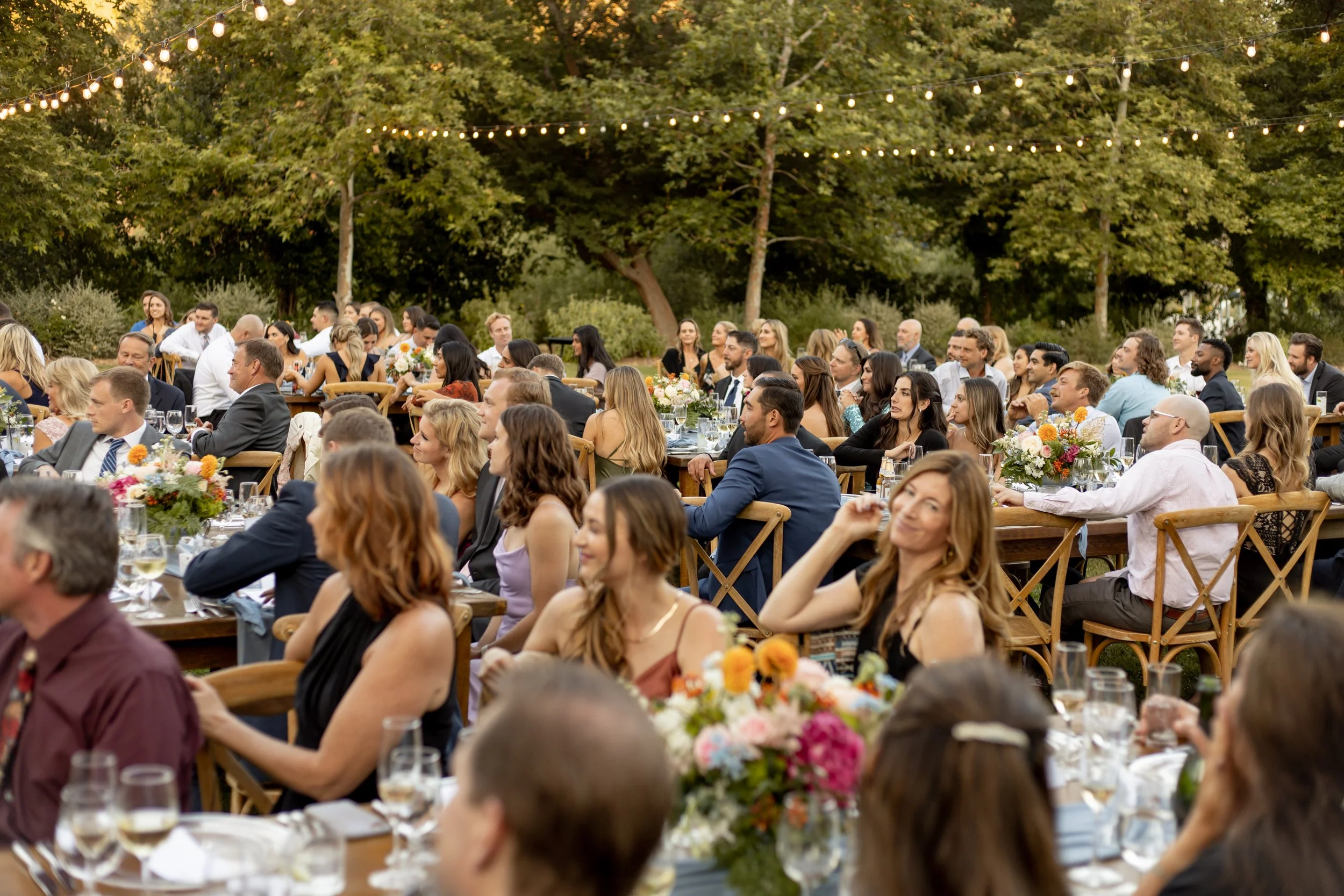 Well-dressed wedding guests are seated at wood tables for outdoor dining on the lawn at The Lodge at Malibou Lake in Agoura Hills, CA.