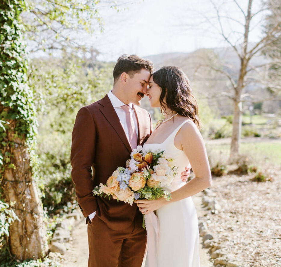 A bride and groom stand closely together outdoors, touching foreheads and smiling, with the bride holding a bouquet of flowers. Spring trees and bright sunlight in the background.