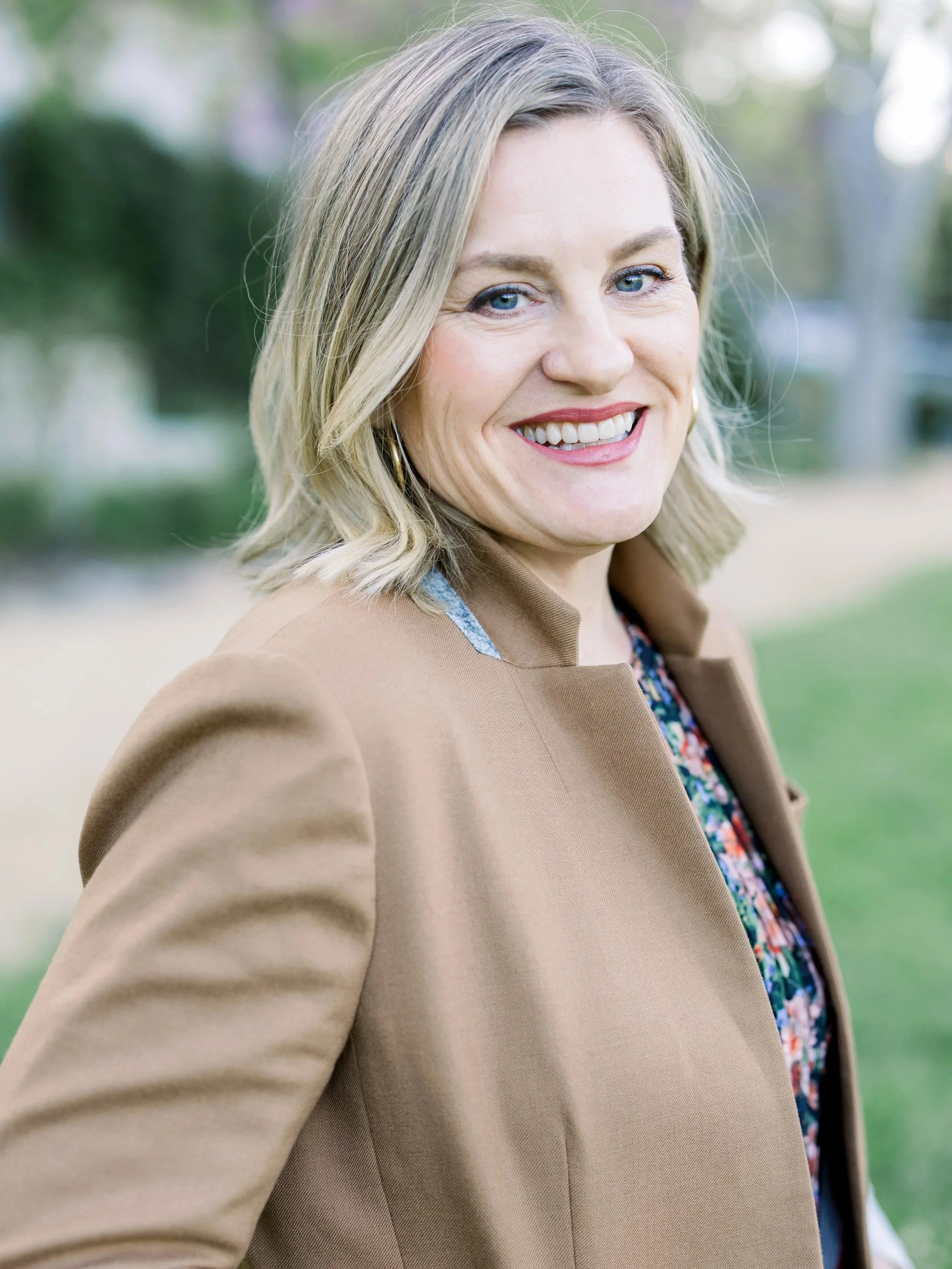 A woman with blonde hair, blue eyes, and a bright smile is outdoors, wearing a beige coat over a floral dress. The background is blurred greenery.