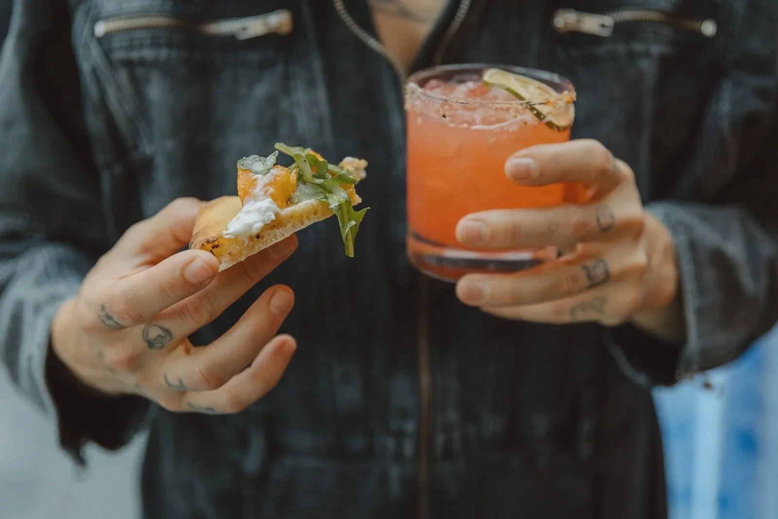 Person wearing a black leather jacket holding a slice of pizza with toppings and a glass of pink-colored drink with lime wedge. Wedding Catering by Chef Cordelia.