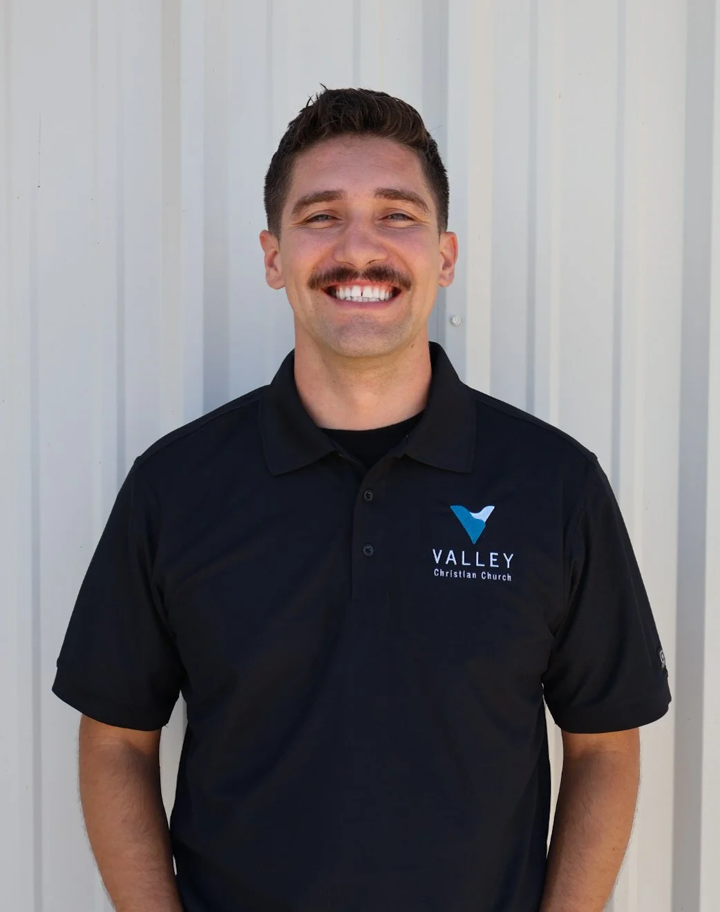 A smiling man with a mustache and short dark hair wearing a black polo shirt with 'Valley Christian Church' logo, standing against a white wall.
