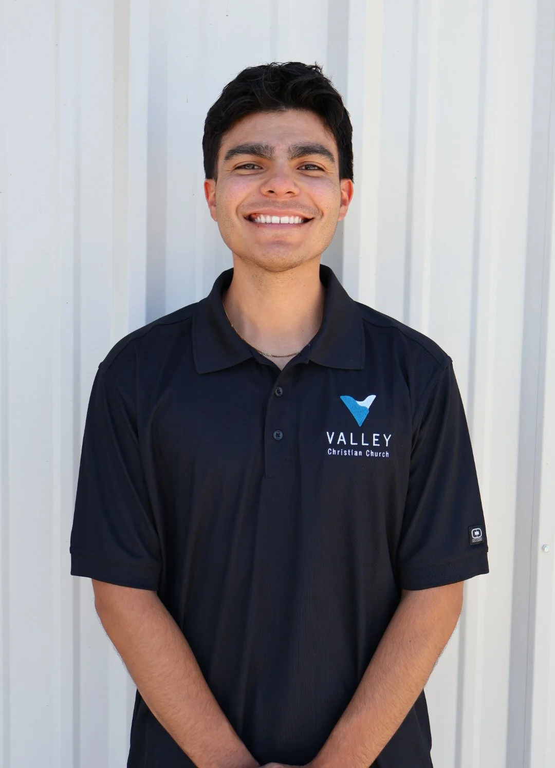 A young man with dark hair, smiling, stands in front of a white metal wall, wearing a black Vally Christian Church polo shirt.