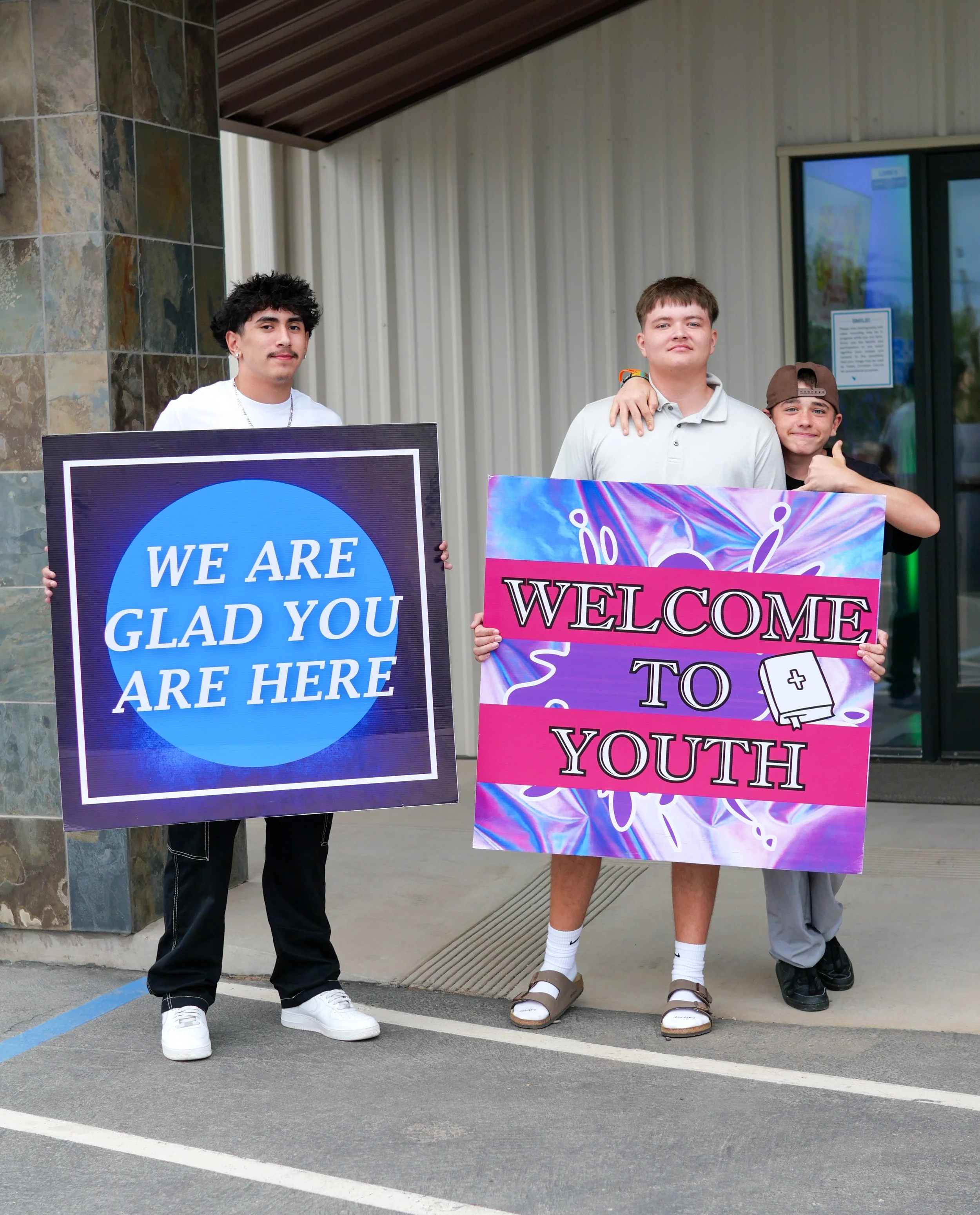 Three young men standing outside a building holding welcoming signs for youth.