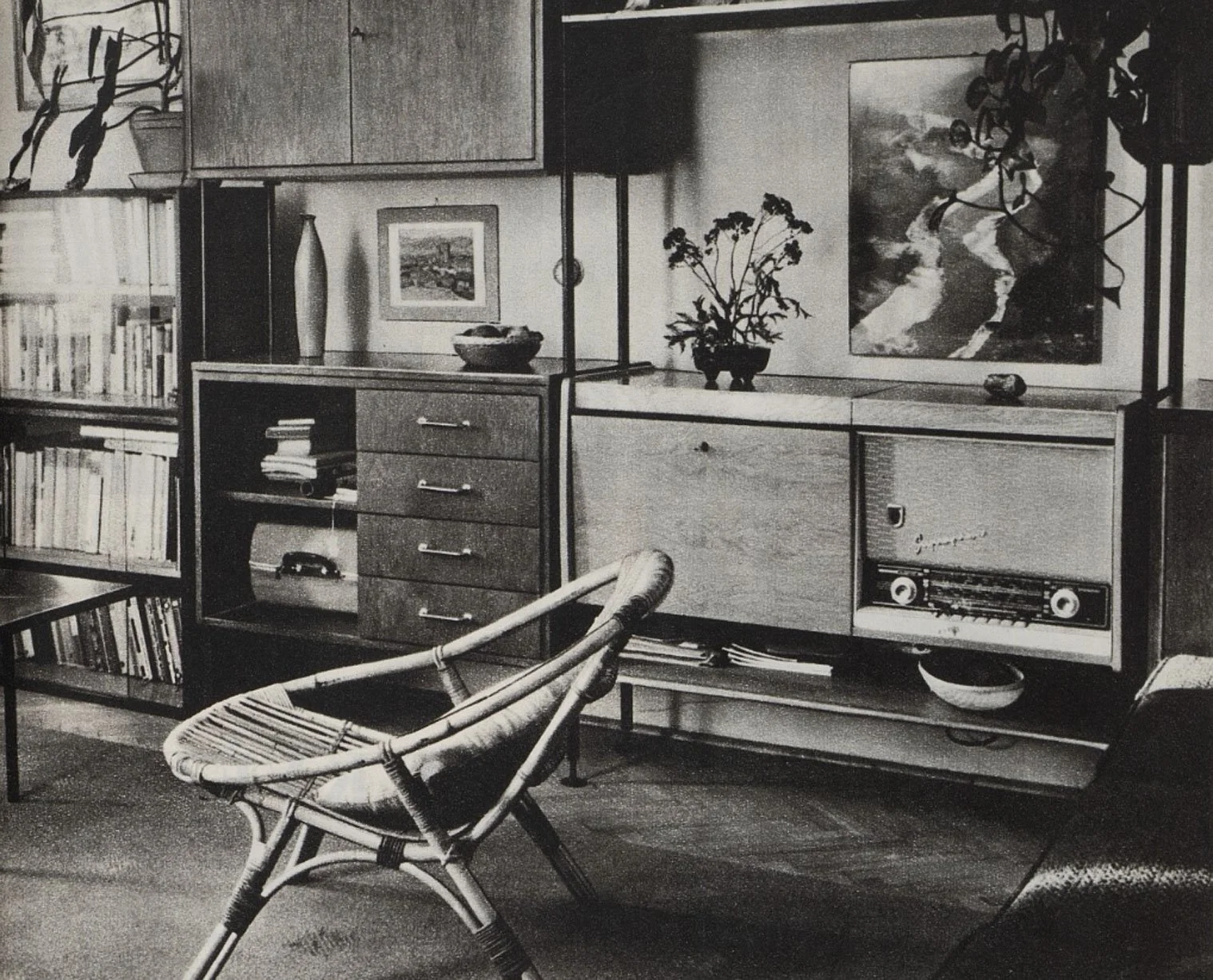 Historical black-and-white interior photograph showing a rattan lounge chair in front of storage furniture
