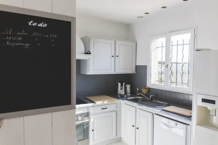 A white kitchen with white cabinets, a black countertop, and a window with metal bars. There are small appliances on the counter, including a coffee maker and a kettle, along with two cutting boards. A chalkboard with a to-do list is visible on the left.