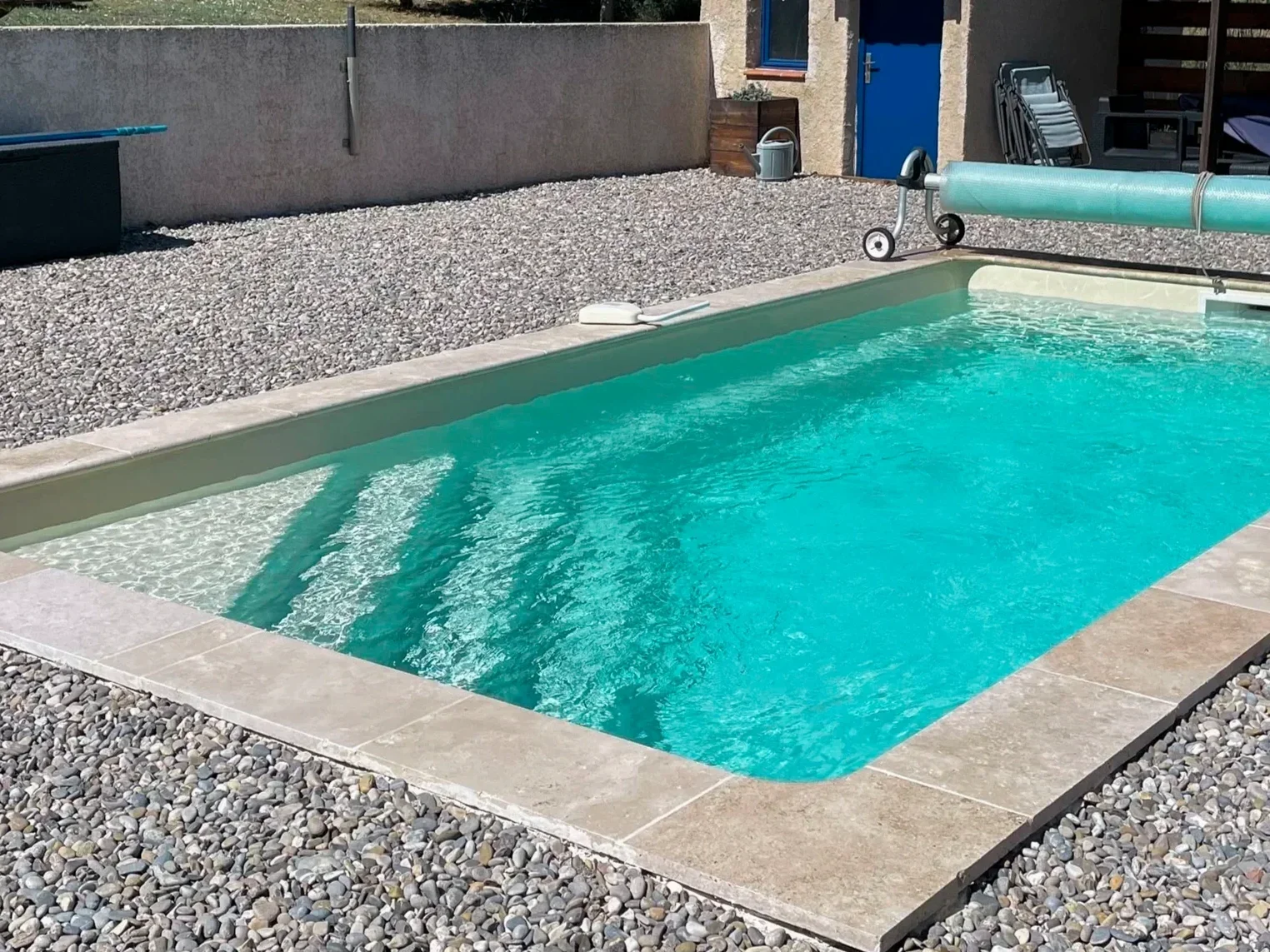 Backyard swimming pool with tile border, surrounded by gray gravel, with a pool cover roller at one end and a rule on the pool edge, with a blue door and various outdoor furniture and storage items in the background.