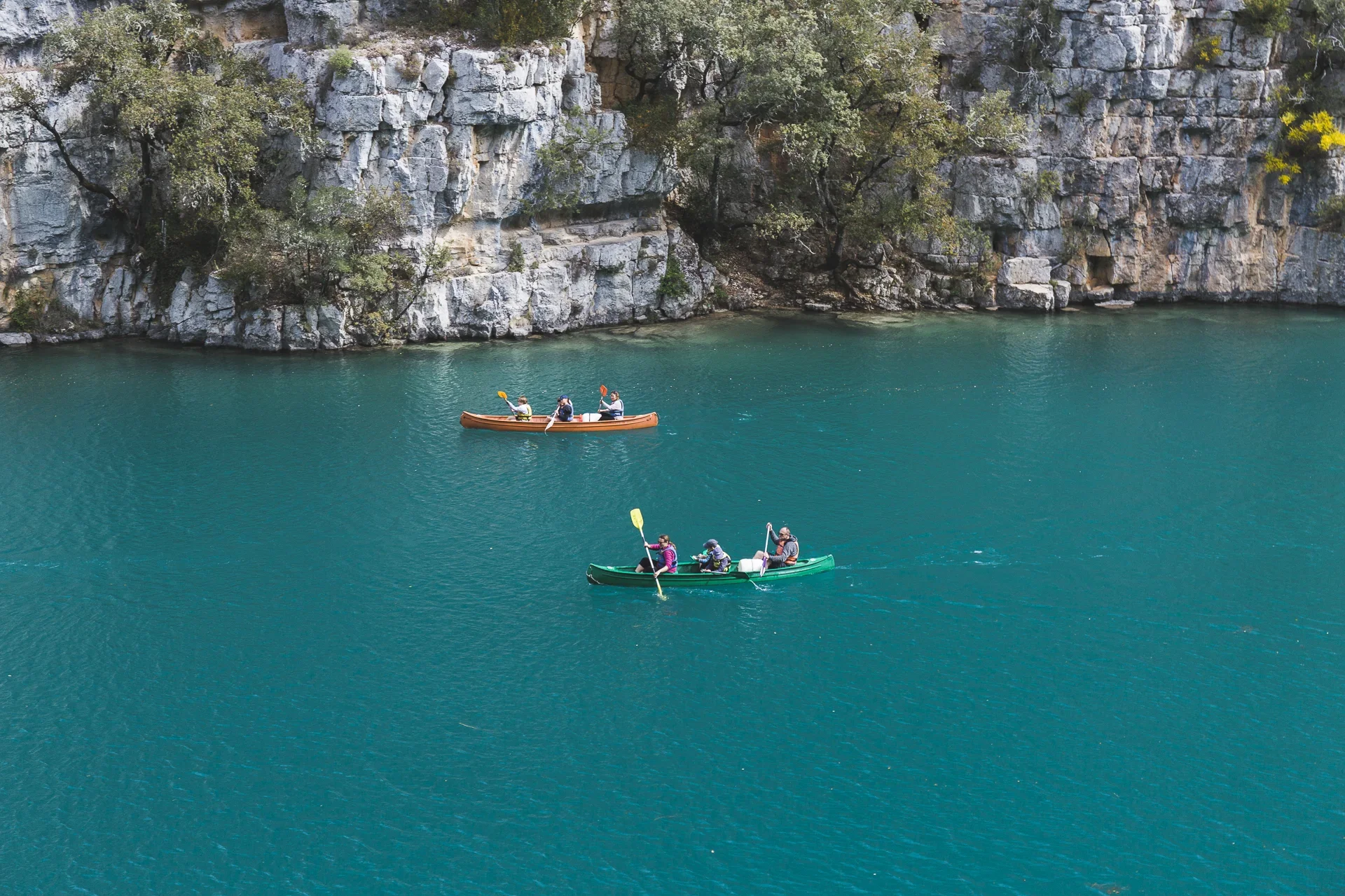 Three kayaks with people paddling on a greenish-blue body of water near a rocky cliff face with trees.