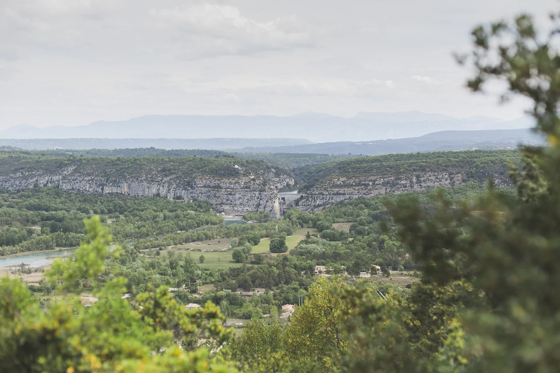 Scenic landscape of green hills, cliffs, a river, and a dam in the distance, with cloudy sky overhead.