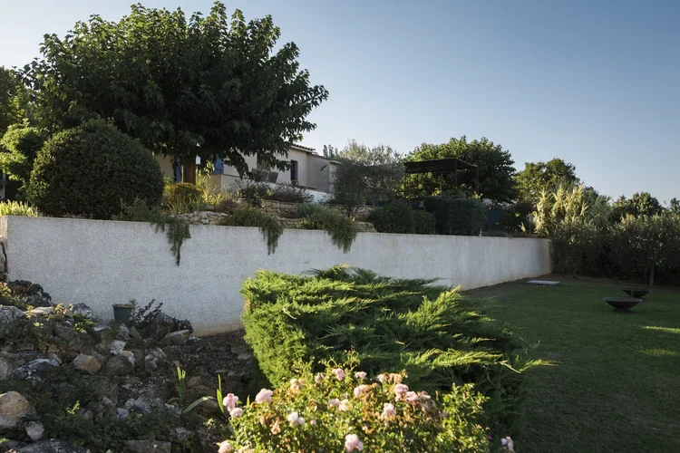 A backyard garden with a white retaining wall, green shrubs, trees, and pink flowers, under a clear blue sky.