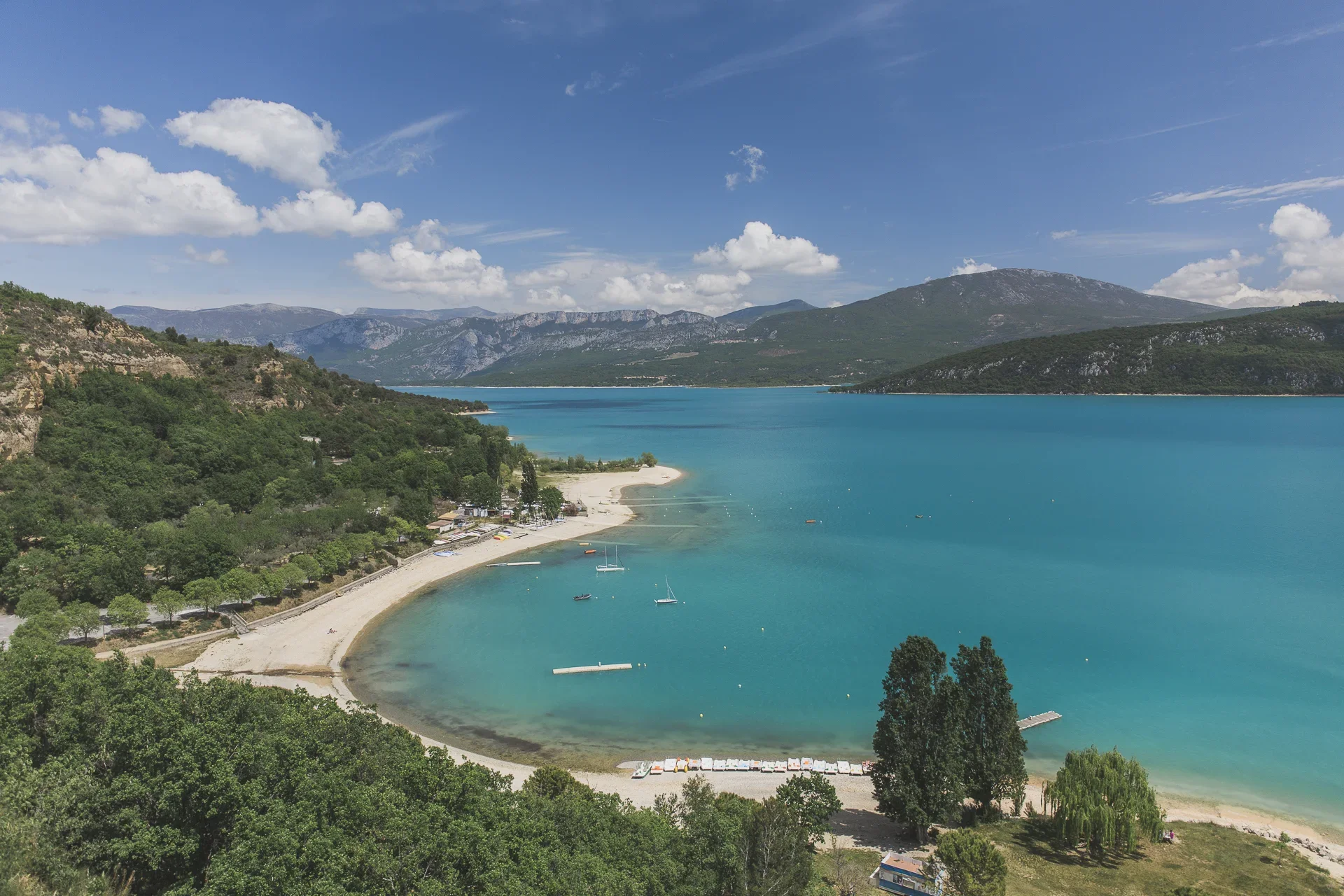 A scenic view of a turquoise lake surrounded by green hills and mountains, with a sandy beach and boats on the water under a partly cloudy sky.