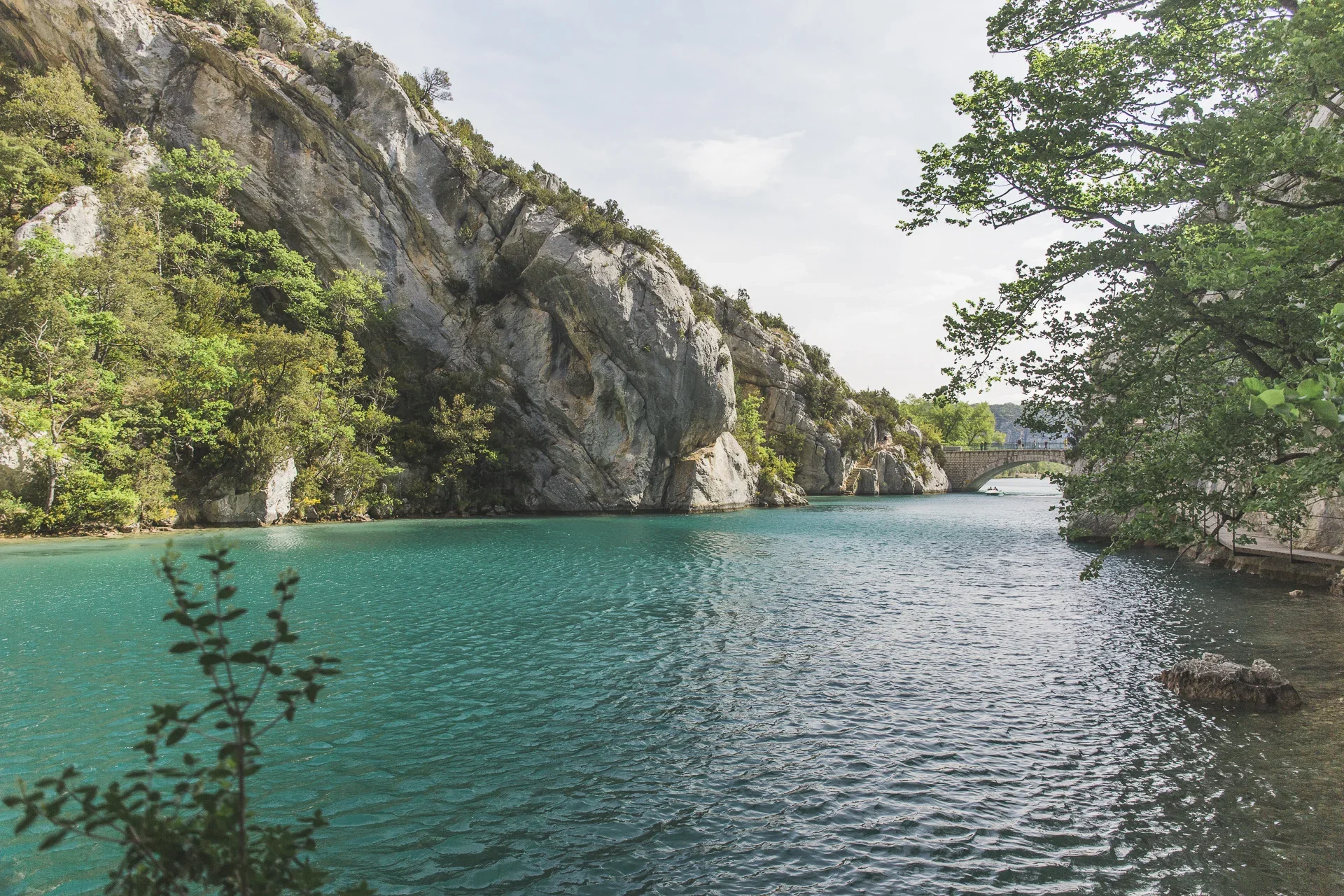 A peaceful river flows between rocky cliffs and green trees, with a bridge visible in the distance on a cloudy day.