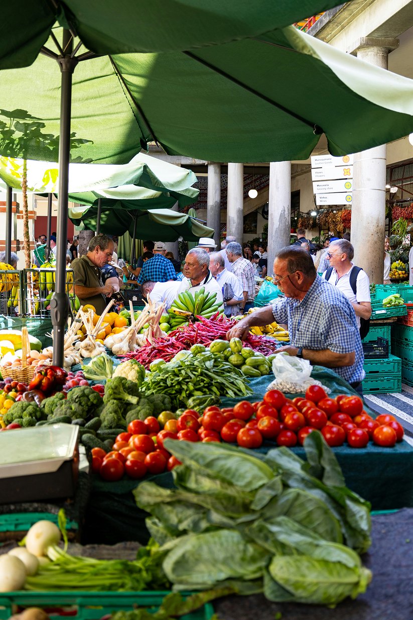 Vendors and shoppers at an outdoor farmers market with baskets of fresh vegetables and fruits under green umbrellas.