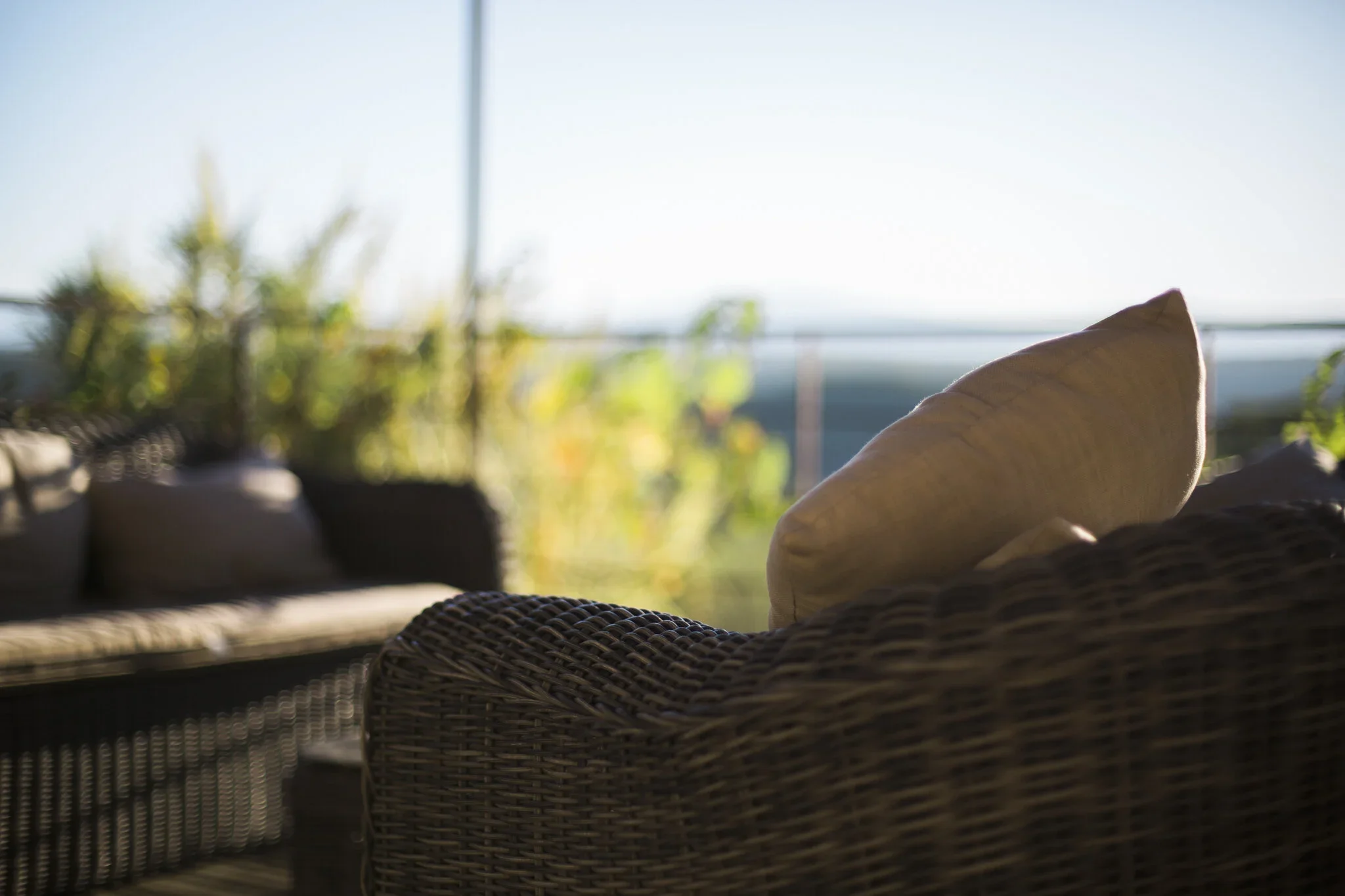 Wicker outdoor furniture with cushions on a porch or patio, overlooking a bright outdoor scene with plants and a clear sky.