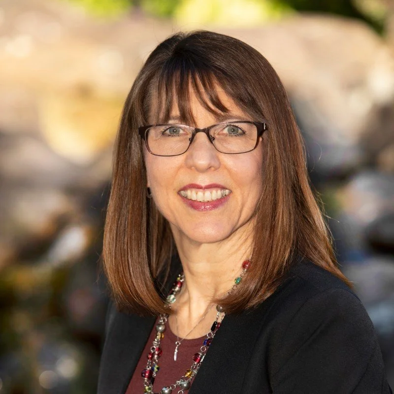 Smiling woman with straight brown hair, glasses, and colorful jewelry, outdoors, blurred background.