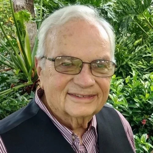 An elderly man with gray hair and glasses smiling outdoors, surrounded by green tropical plants.
