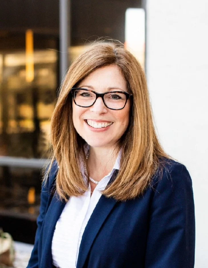 A woman with shoulder-length reddish-brown hair, glasses, and a navy blazer smiling in an office setting.