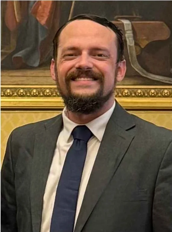 Smiling man in a gray suit with a dark tie, standing in front of a gold-framed painting.