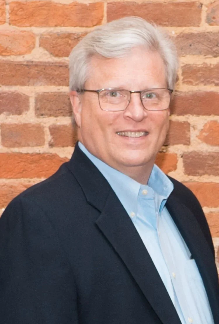 Portrait of a man with gray hair and glasses, wearing a dark blazer and light blue shirt, standing against a brick wall background.