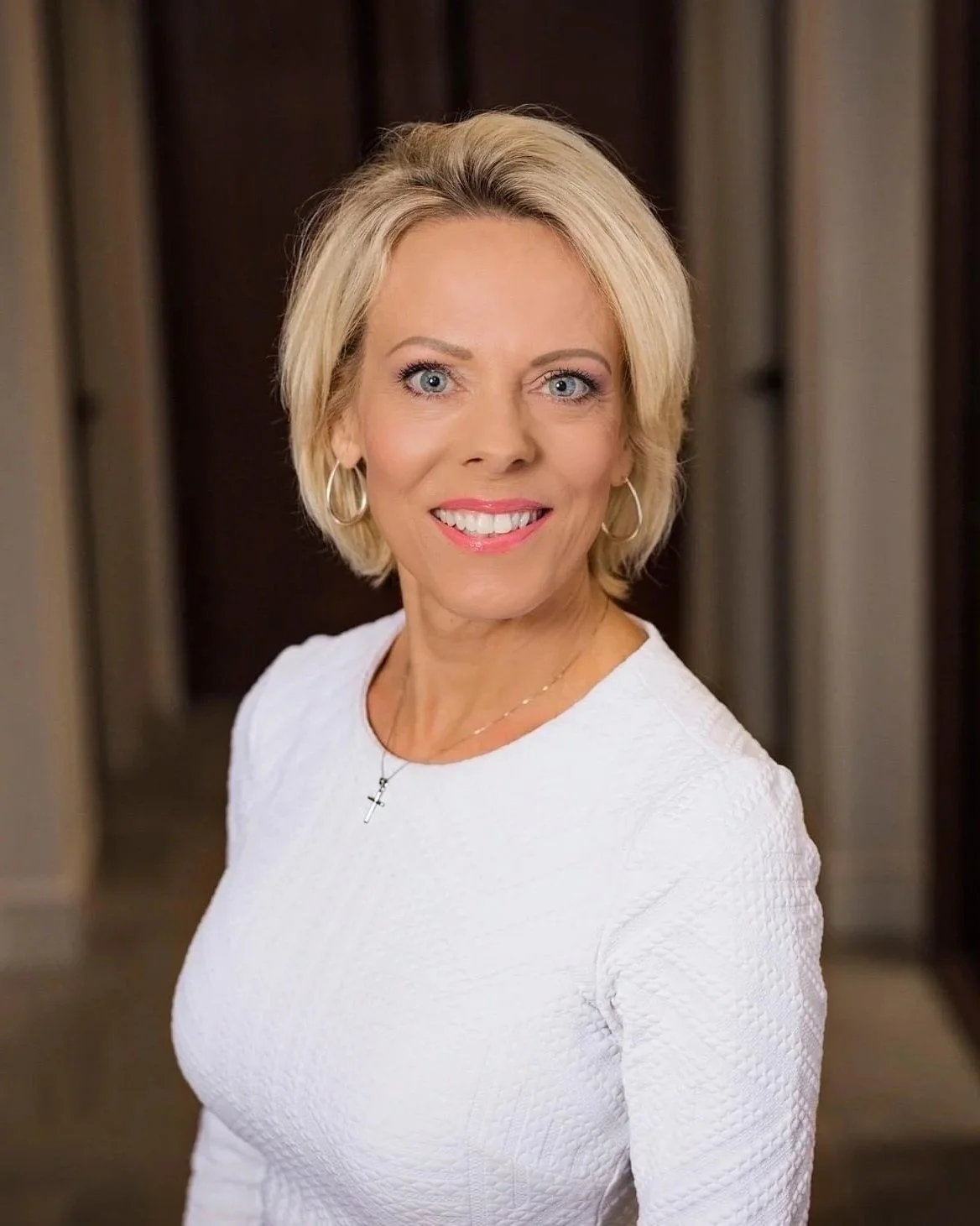Portrait of a smiling middle-aged woman with blonde hair, wearing a white top, hoop earrings, a cross necklace, and makeup, standing indoors with a blurred background.
