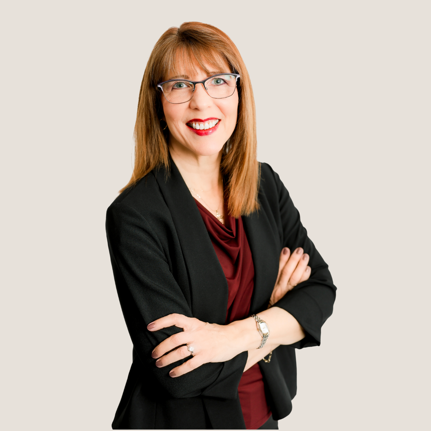Professional woman with red hair and glasses smiling, wearing a black blazer and burgundy top, standing with arms crossed against a neutral background.