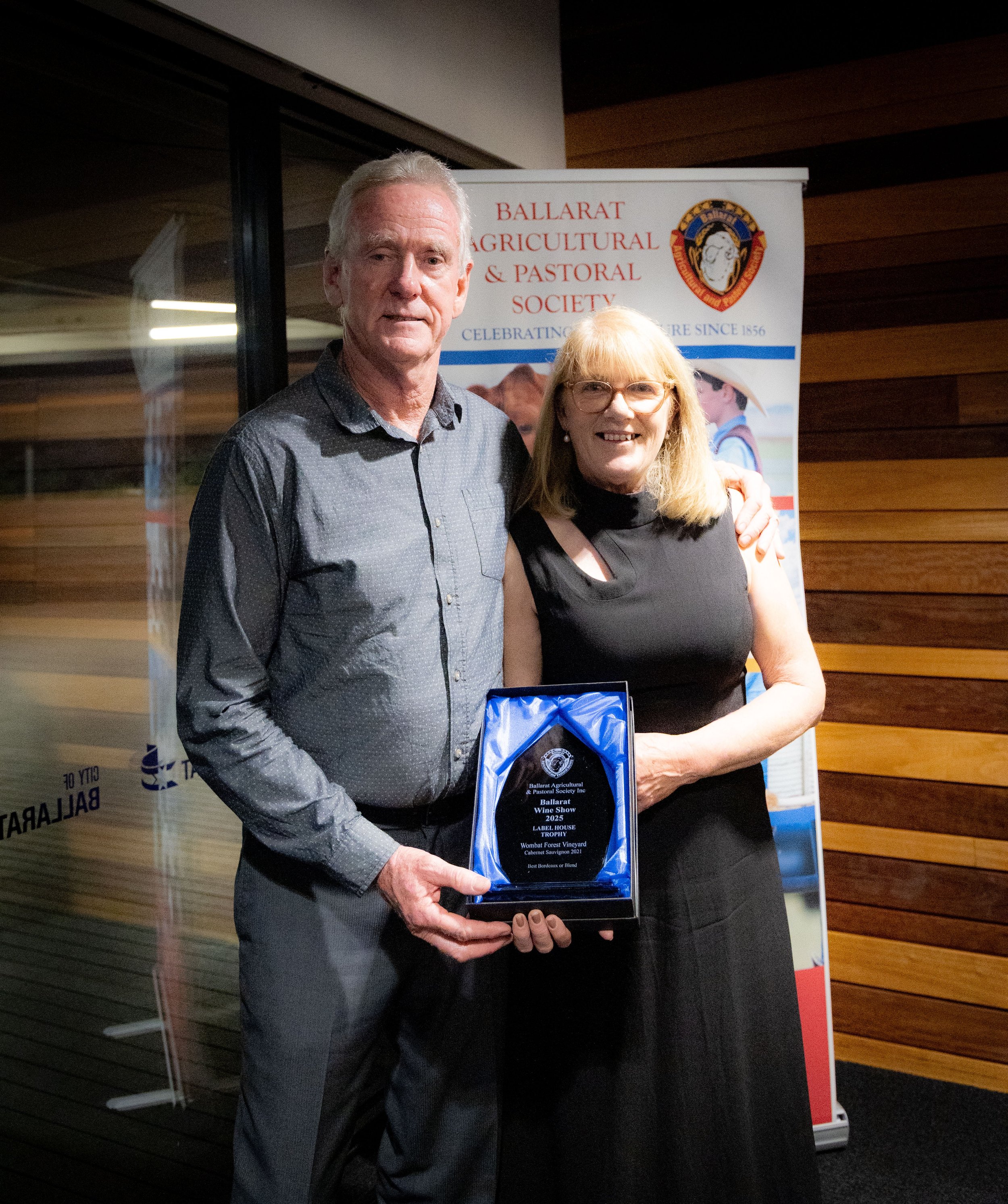A man and woman standing together holding an award plaque at an event for the Ballarat Agricultural & Pastoral Society, with a banner behind them displaying the society's name and logo.